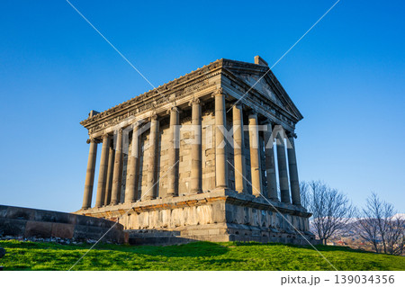 Sunlit ancient Garni temple with stone columns, grassy hill, and bare trees 139034356