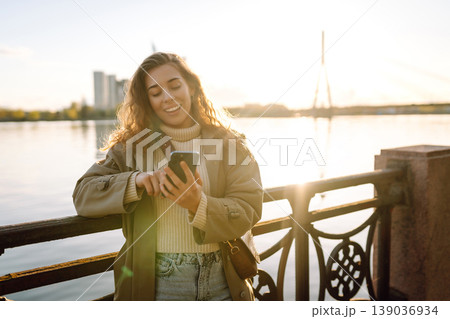 Young woman enjoys a sunny day by the river, using her smartphone while smiling in the warm sunlight Young woman enjoys a sunny day by the river, using her smartphone while smiling in the warm sunlight 139036934