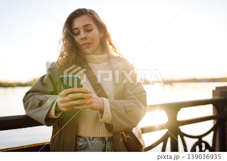 Young female city dweller with mobile phone standing on embankment alone. Woman shopping online. Young female city dweller with mobile phone standing on embankment alone. Woman shopping online. 139036935