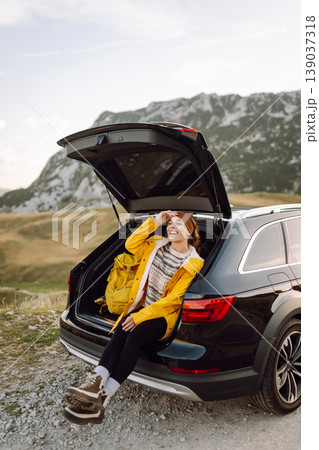 A cheerful traveler enjoying a scenic mountain view while sitting on the trunk. Travelling by car. A cheerful traveler enjoying a scenic mountain view while sitting on the trunk. Travelling by car. 139037318