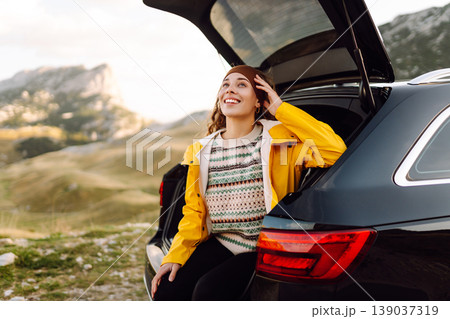 A cheerful traveler enjoying a scenic mountain view while sitting on the trunk. Travelling by car. A cheerful traveler enjoying a scenic mountain view while sitting on the trunk. Travelling by car. 139037319
