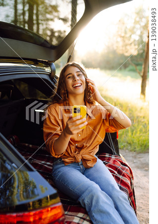 Young curly woman in stylish clothes in the trunk of a car in the forest and uses the phone. Young curly woman in stylish clothes in the trunk of a car in the forest and uses the phone. 139038493