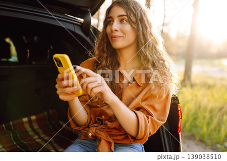 Young curly woman in stylish clothes in the trunk of a car in the forest and uses the phone. 139038510