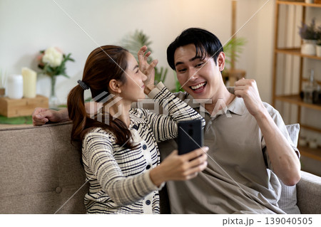 Asian adult couple viewing smartphone screen expressing happiness sharing good news winning exciting content home living room comfortable sofa together Asian adult couple viewing smartphone screen expressing happiness sharing good news winning exciting content home living room comfortable sofa together 139040505