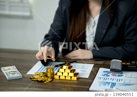 Asian adult woman investor manages financial wealth gold coins bars cash for business strategy and growth at office desk 139040551