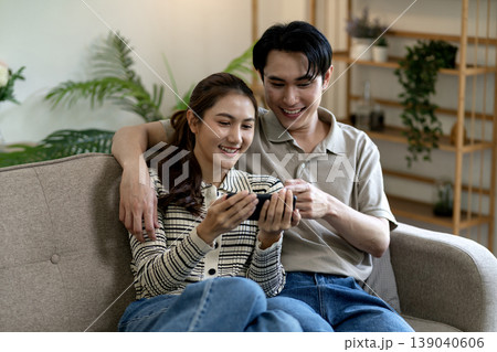 Asian young adult couple sitting on sofa happily watching smartphone enjoying digital content together home lifestyle Asian young adult couple sitting on sofa happily watching smartphone enjoying digital content together home lifestyle 139040606