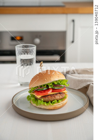 Beef burger with tomato, cheese, and lettuce on a plate on a light table with glass of water Beef burger with tomato, cheese, and lettuce on a plate on a light table with glass of water 139040712
