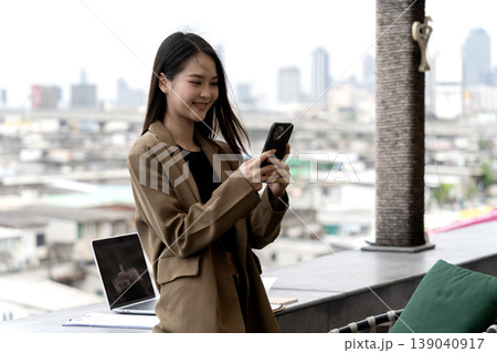 Asian adult woman professional uses smartphone for digital communication connection engaging mobile technology enjoying vibrant city rooftop view urban modern work life 139040917