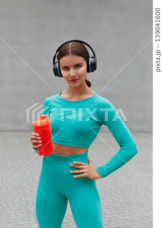 Beautiful female runner standing outdoors holding water bottle and using wireless headphones. Fitness woman taking a break after running workout. Beautiful female runner standing outdoors holding water bottle and using wireless headphones. Fitness woman taking a break after running workout. 139041600
