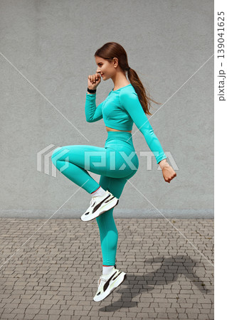 Young woman with fit body jumping and running against grey background. Female model in blue sportswear exercising outdoors Young woman with fit body jumping and running against grey background. Female model in blue sportswear exercising outdoors 139041625