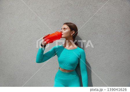 Beautiful girl runner standing outdoors, drinking water of bottle. Fitness woman taking a break after running workout. Beautiful girl runner standing outdoors, drinking water of bottle. Fitness woman taking a break after running workout. 139041626