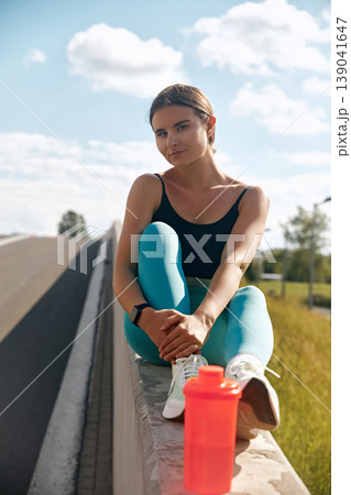 Fit woman in athletic wear sitting on a ledge beside a road, with a water bottle nearby, enjoying a moment of relaxation after an intense workout session outdoors. Fit woman in athletic wear sitting on a ledge beside a road, with a water bottle nearby, enjoying a moment of relaxation after an intense workout session outdoors. 139041647