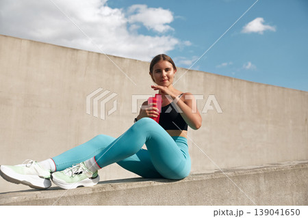 Fit young woman in stylish activewear sits on a concrete ledge, holding a refreshing drink, showcasing a sporty lifestyle and vibrant energy in a modern outdoor setting. Fit young woman in stylish activewear sits on a concrete ledge, holding a refreshing drink, showcasing a sporty lifestyle and vibrant energy in a modern outdoor setting. 139041650