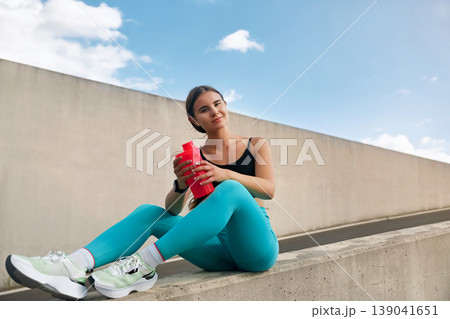 Fit woman in athletic wear sits on a concrete barrier, holding a red water bottle, with a bright blue sky and clouds in the background, embodying a sporty and active lifestyle. 139041651
