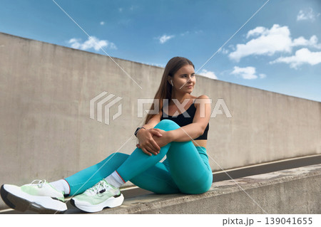 Fit woman in turquoise leggings and black crop top sits on a concrete wall, enjoying a moment of relaxation after workout, with a vibrant orange water bottle beside her. Fit woman in turquoise leggings and black crop top sits on a concrete wall, enjoying a moment of relaxation after workout, with a vibrant orange water bottle beside her. 139041655