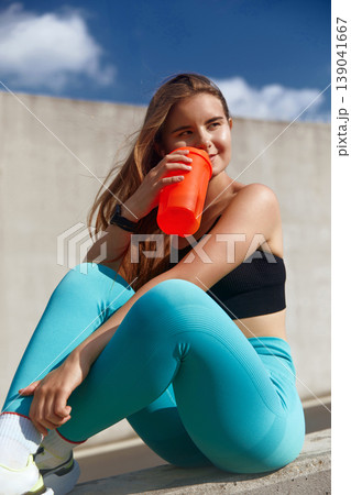 Young woman dressed in stylish athletic clothing sits comfortably on a ledge, sipping a vibrant orange sports drink, embodying a casual fitness lifestyle in a bright outdoor setting with clouds. 139041667