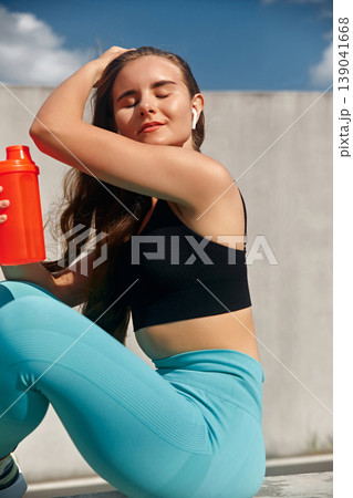 Young woman with long hair dressed in a black sports top and blue leggings sits outdoors, holding a vibrant orange shaker bottle, radiating fitness and vitality in a bright, sunny atmosphere. Young woman with long hair dressed in a black sports top and blue leggings sits outdoors, holding a vibrant orange shaker bottle, radiating fitness and vitality in a bright, sunny atmosphere. 139041668