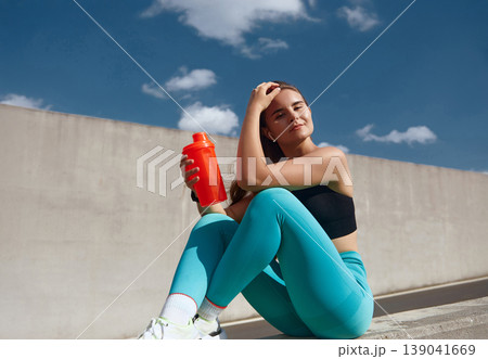 Athletic woman dressed in black crop top and turquoise leggings sits on a ledge, holding an orange shaker bottle, exuding confidence and vitality after an intense workout session. Athletic woman dressed in black crop top and turquoise leggings sits on a ledge, holding an orange shaker bottle, exuding confidence and vitality after an intense workout session. 139041669