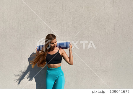 Fit young woman with long hair wearing a black sports bra and turquoise leggings carries a yoga mat over her shoulder against a textured concrete wall, embodying a healthy lifestyle. Fit young woman with long hair wearing a black sports bra and turquoise leggings carries a yoga mat over her shoulder against a textured concrete wall, embodying a healthy lifestyle. 139041675