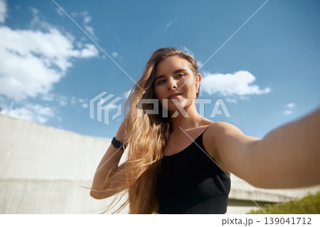 Fit young woman with long hair taking a selfie outdoors, showcasing a sporty outfit against a bright blue sky, capturing the essence of fitness and self-expression in an active lifestyle. Fit young woman with long hair taking a selfie outdoors, showcasing a sporty outfit against a bright blue sky, capturing the essence of fitness and self-expression in an active lifestyle. 139041712