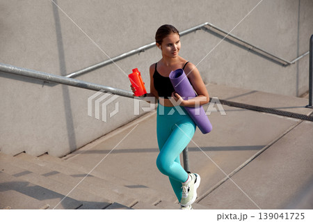 Fit woman in activewear holds a water bottle and yoga mat while leaning against a railing, showcasing a sporty lifestyle and dedication to fitness and wellness in an urban environment. Fit woman in activewear holds a water bottle and yoga mat while leaning against a railing, showcasing a sporty lifestyle and dedication to fitness and wellness in an urban environment. 139041725