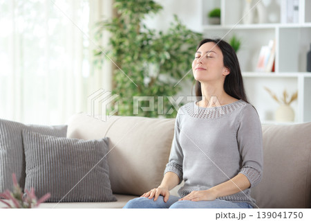 Asian woman breathing fresh air seated on a couch at home Asian woman breathing fresh air seated on a couch at home 139041750