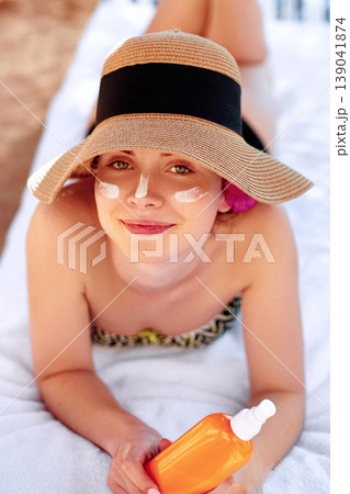 Young woman with sun cream on face holding sunscren bottle on the beach. Female in hat applying moisturizing lotion on skin. Young woman with sun cream on face holding sunscren bottle on the beach. Female in hat applying moisturizing lotion on skin. 139041874