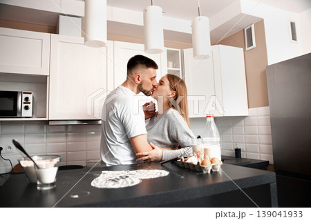 Couple shares a kiss in a modern kitchen while preparing breakfast together in the morning light with smiles and warmth in the air Couple shares a kiss in a modern kitchen while preparing breakfast together in the morning light with smiles and warmth in the air 139041933