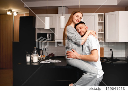 Couple enjoy time together in kitchen while sitting on counter during morning routine in modern home setting Couple enjoy time together in kitchen while sitting on counter during morning routine in modern home setting 139041938