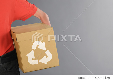 A person holding a brown cardboard box featuring a white recycling logo against a neutral gray background, representing environmental sustainability and waste reduction A person holding a brown cardboard box featuring a white recycling logo against a neutral gray background, representing environmental sustainability and waste reduction 139042126