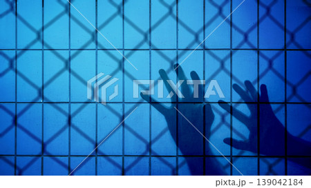 A silhouette of hands reaching out against a blue tiled wall with a chain-link fence shadow, representing imprisonment, confinement, isolation, and the struggle for freedom 139042184