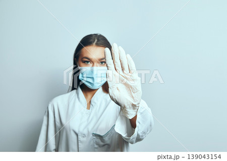 Medical professional wearing gloves and a mask gestures to stop while standing against a plain background 139043154