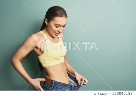 Woman shows her success in weight loss by fitting into larger jeans while standing against a simple background in a room Woman shows her success in weight loss by fitting into larger jeans while standing against a simple background in a room 139043158