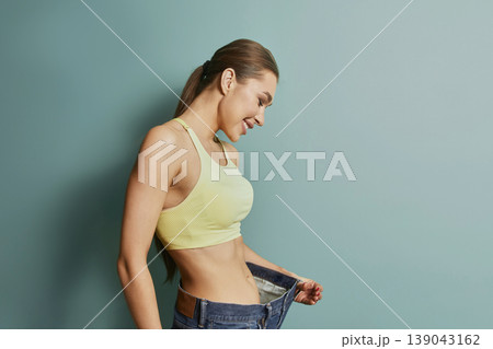 Woman smiles while holding oversized pants in front of green wall during indoor activity showcasing weight loss achievement 139043162