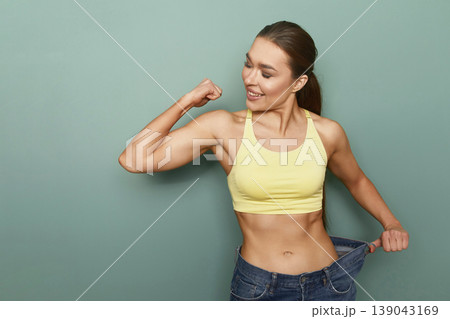 Woman shows off fitness progress while flexing arm muscles in casual outfit against a plain green background during daylight hours Woman shows off fitness progress while flexing arm muscles in casual outfit against a plain green background during daylight hours 139043169