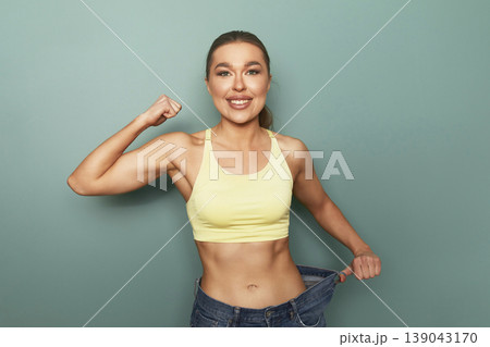 Woman shows weight loss success standing against green wall wearing yellow top and blue jeans after fitness journey and healthy changes in lifestyle Woman shows weight loss success standing against green wall wearing yellow top and blue jeans after fitness journey and healthy changes in lifestyle 139043170