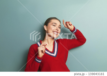 Woman enjoys music while dancing in a red sweater against a blue wall during daytime Woman enjoys music while dancing in a red sweater against a blue wall during daytime 139043181