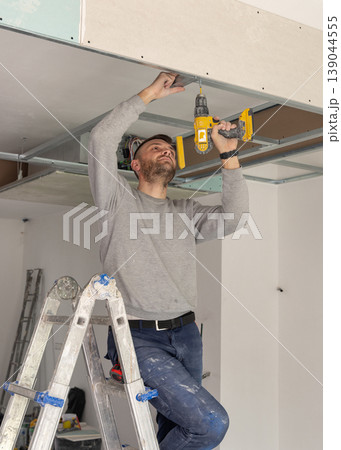 Construction worker using drill on metal frame for drywall suspended ceiling in apartment renovation 139044555