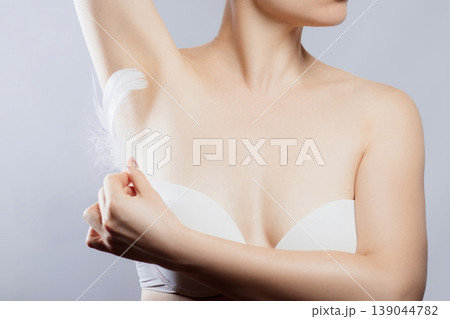 Woman applying lotion to underarm area while wearing a white top in a bright indoor setting 139044782