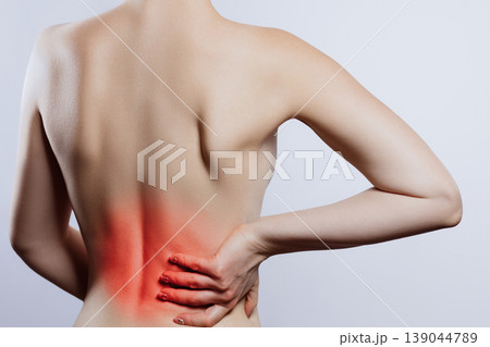 Woman holds her lower back in pain while standing against a plain background Woman holds her lower back in pain while standing against a plain background 139044789