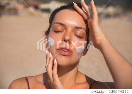 Young Woman Applying Sunscreen On Face With Eyes Closed At Beach. Beauty Skincare Concept. Female Using Sun Cream For Skin Protection Young Woman Applying Sunscreen On Face With Eyes Closed At Beach. Beauty Skincare Concept. Female Using Sun Cream For Skin Protection 139044832