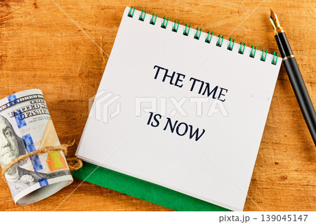Rolled US 100 dollar bills tied with twine sit on a wooden surface next to a notebook displaying THE TIME IS NOW and a fountain pen 139045147