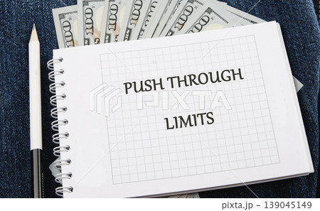White spiral notebook featuring PUSH THROUGH LIMITS on grid paper rests on blue denim. One hundred US Dollar bills and a white pencil are nearby White spiral notebook featuring PUSH THROUGH LIMITS on grid paper rests on blue denim. One hundred US Dollar bills and a white pencil are nearby 139045149
