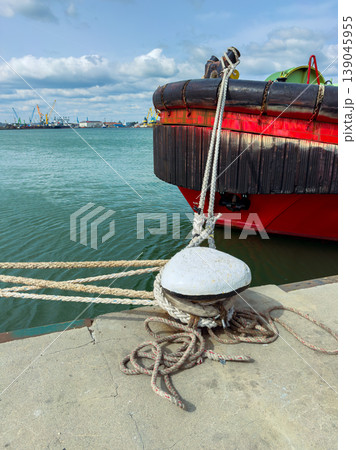 Rope tied to cleat on dock with boat floating in water during daytime near harbor with cranes and ships in the background Rope tied to cleat on dock with boat floating in water during daytime near harbor with cranes and ships in the background 139045955