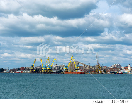 View of a busy port with cranes and ships working during the day near a city skyline with clouds in the sky View of a busy port with cranes and ships working during the day near a city skyline with clouds in the sky 139045958
