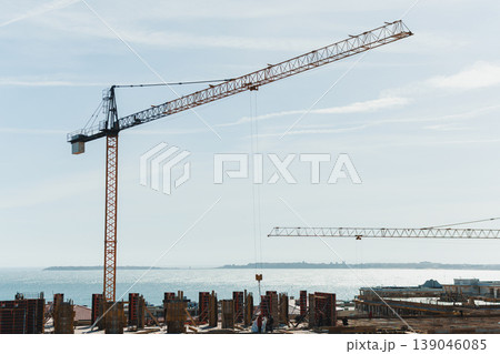 Construction work takes place by the water's edge with cranes, buildings, and workers under a clear sky during the day in an urban area 139046085