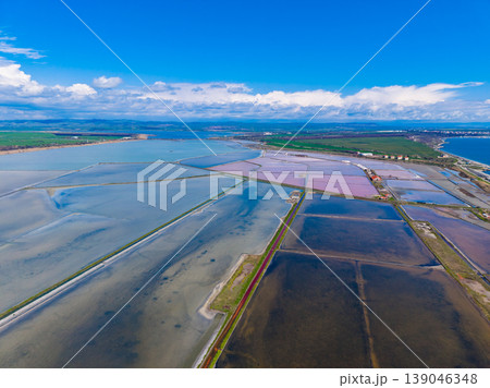 Salt fields stretch across the landscape near the coast, showing clear water and patches of pink under a bright blue sky in the daytime 139046348