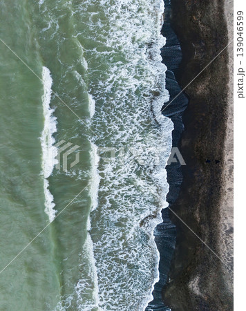 Waves crash onto a shoreline at a beach during daylight hours in a coastal area with dark sand and foamy water 139046599