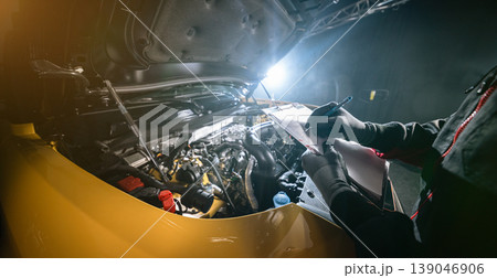 Mechanic inspecting car engine and writing notes on clipboard in workshop. Automotive diagnostics, maintenance checklist and professional service concept. 139046906