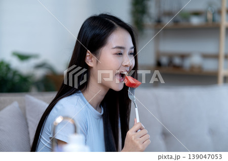 Asian young adult woman enjoying healthy watermelon snack on sofa refreshing fruit indoor bright natural light promoting healthy eating lifestyle wellbeing 139047053